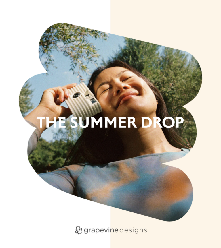 A woman smiling outdoors holds a retro camera to her face under a sunny sky, with “THE SUMMER DROP” text overlaid. Framed by a soft, abstract shape, the image showcases summer promotional products and features the Grapevine Designs logo at the bottom.