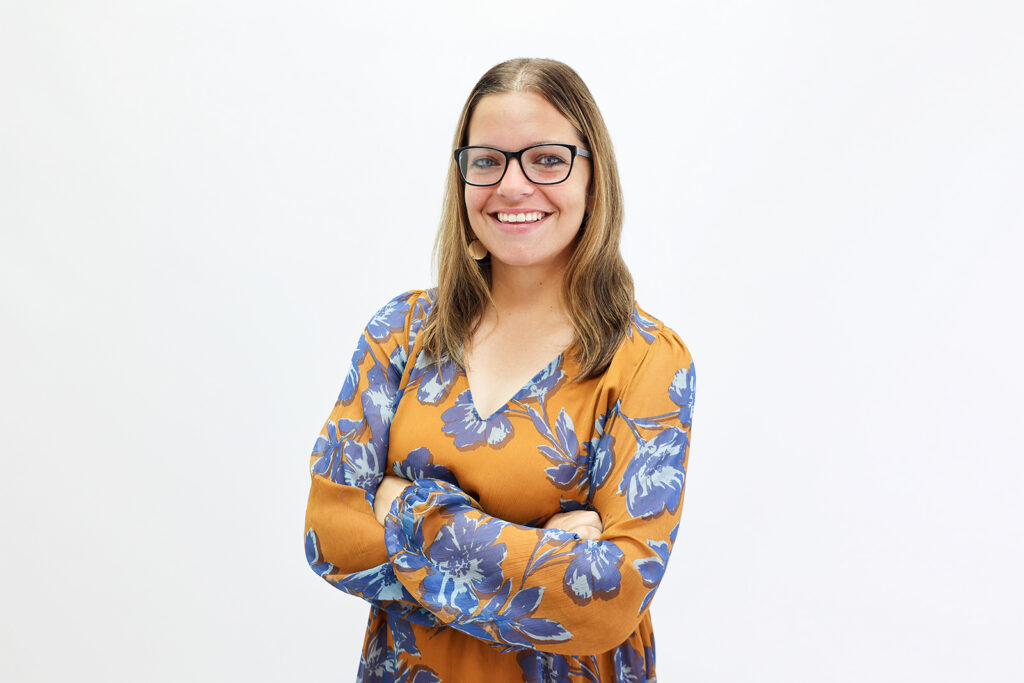 Smiling woman with glasses and brown hair, wearing a yellow dress with blue floral patterns, stands with arms crossed against a plain white background.