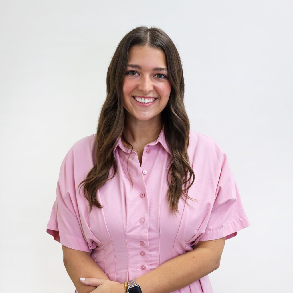 A woman with long brown hair and a pink button-up shirt smiles while standing in front of a plain white background.