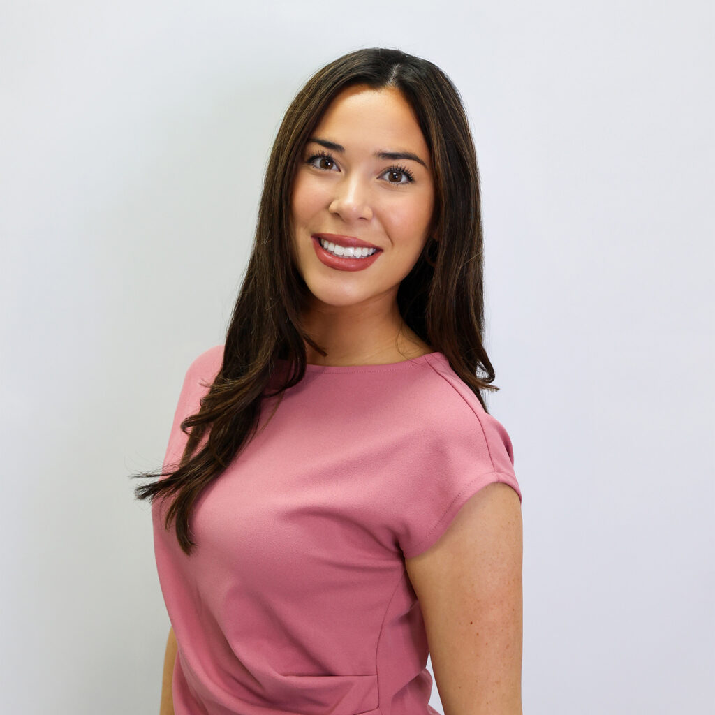 A woman with long brown hair wearing a pink top stands against a plain white background, smiling at the camera.