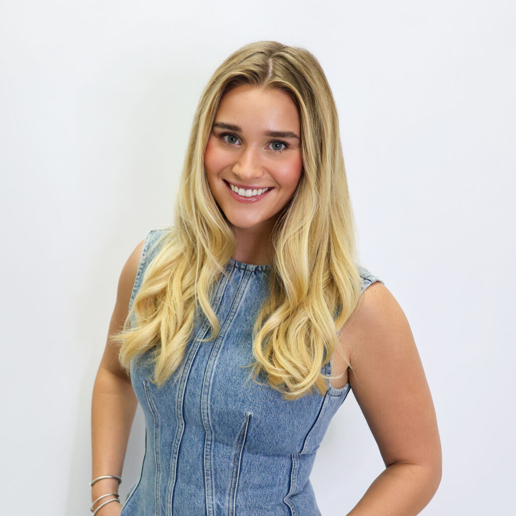 A woman with long blonde hair wearing a sleeveless denim dress smiles at the camera against a plain white background.