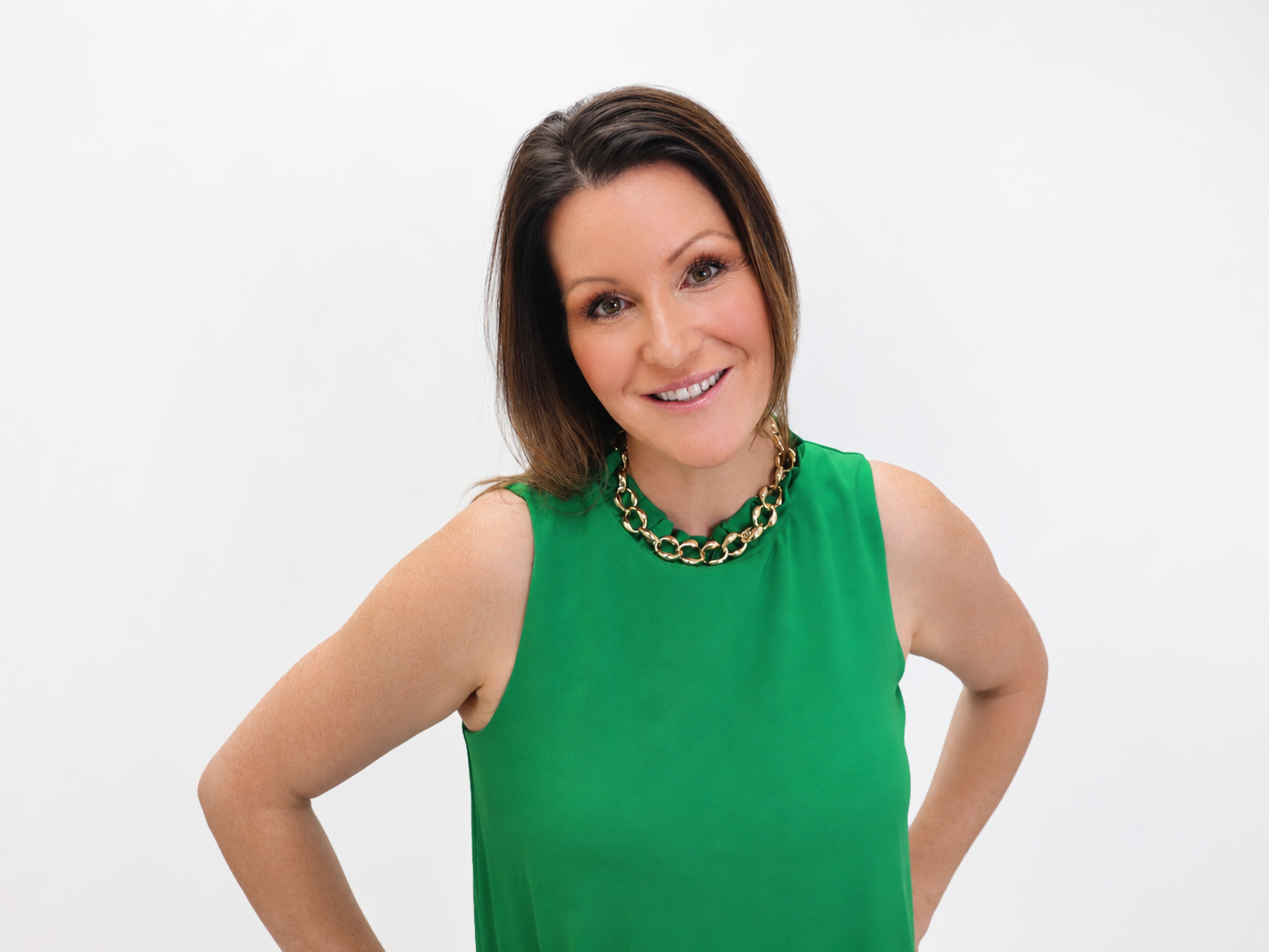 Woman with long brown hair wearing a sleeveless green top and a chunky gold necklace, smiling with her hands on her hips against a plain white background—perfect for representing a confident team member of a promotional marketing company.