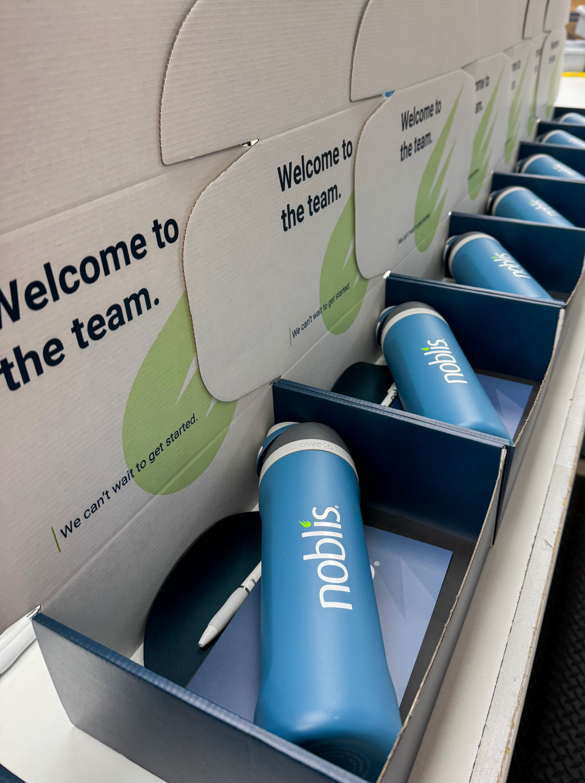 Rows of welcome kits, expertly assembled through kitting, feature blue Noblis water bottles, white pens, and greeting cards arranged in boxes that read “Welcome to the team. We can’t wait to get started.”.