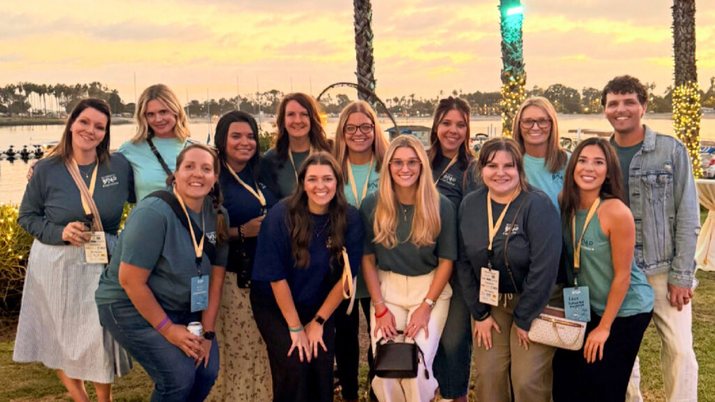 A group of 15 people pose together outdoors at sunset, smiling. They wear teal event shirts and name badges, standing in front of palm trees and lights, with water and sailboats in the background.