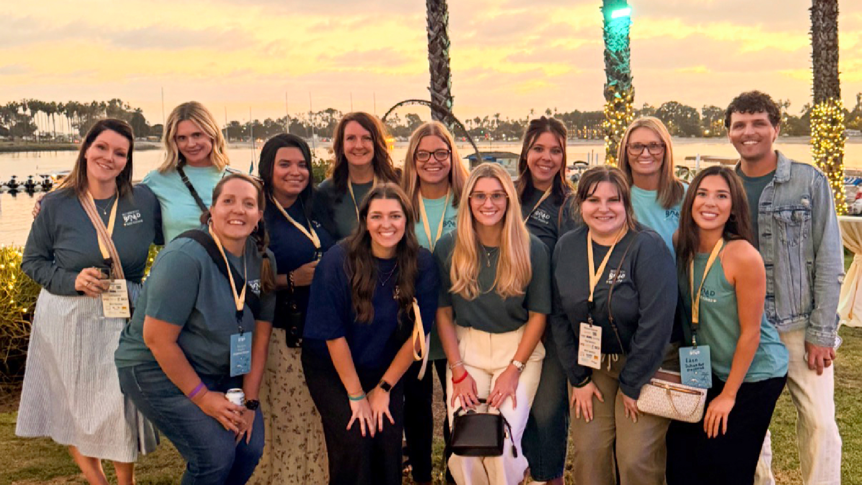A group of 15 people pose together outdoors at sunset, smiling. They wear teal event shirts and name badges, standing in front of palm trees and lights, with water and sailboats in the background.