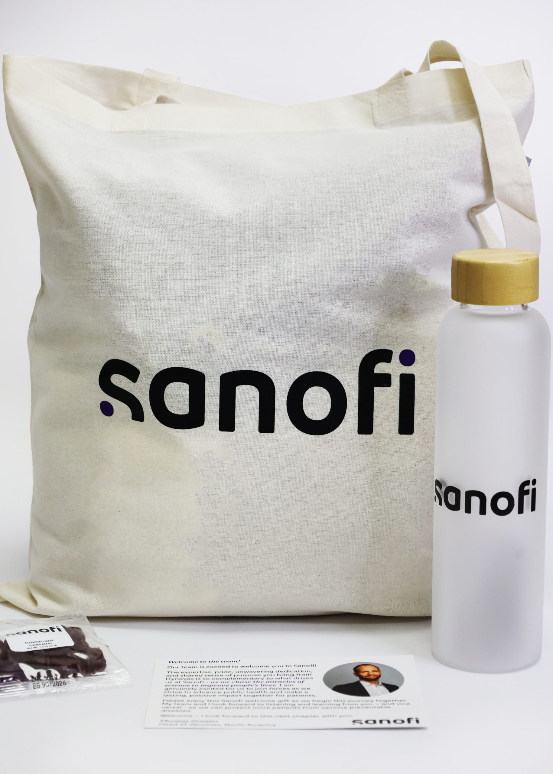 A Sanofi-branded tote bag, a clear water bottle with a tan cap, a wrapped snack, and an informational card—expertly curated by a leading Promotional Marketing Agency—are arranged on a white surface.